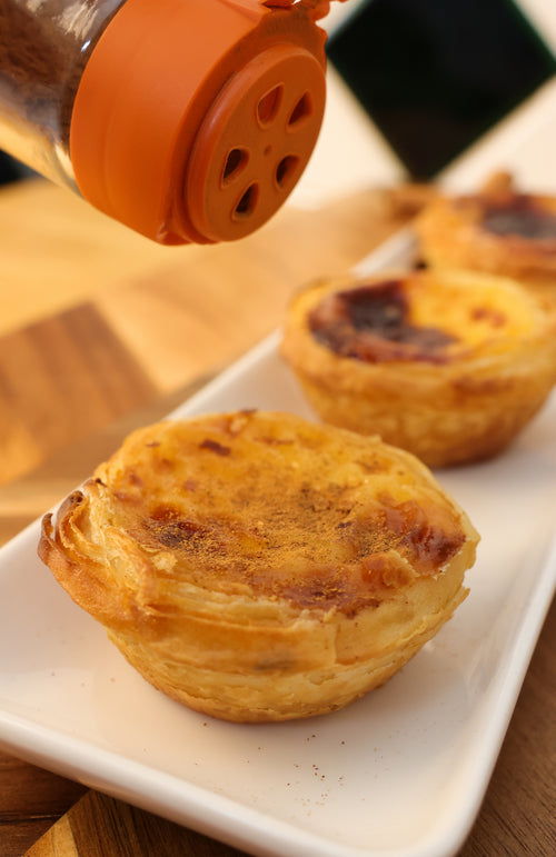 Close-up of baked Portuguese custard tarts or Pastéis de Nata served fresh from the oven with a dusting of cinnamon and powdered sugar next to an espresso coffee.